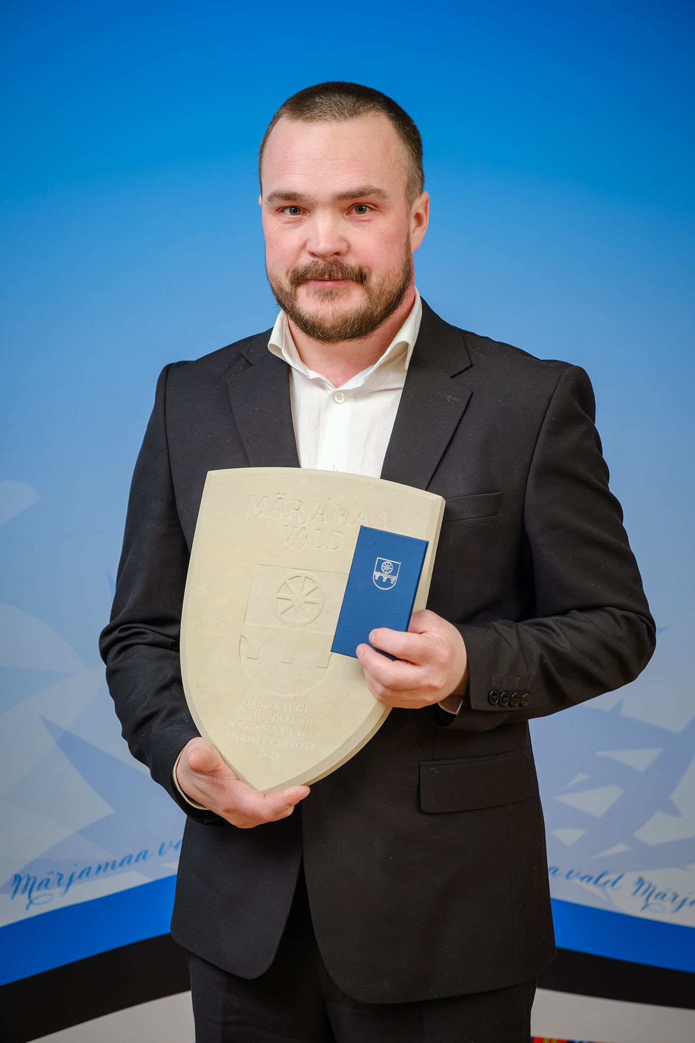 Lühikeste tumedate juustega noor mees, kellel on vuntsid ja lühike habe, hoiab kahe käega enda ees dolomiidist vapikujulist teenetemärki. 