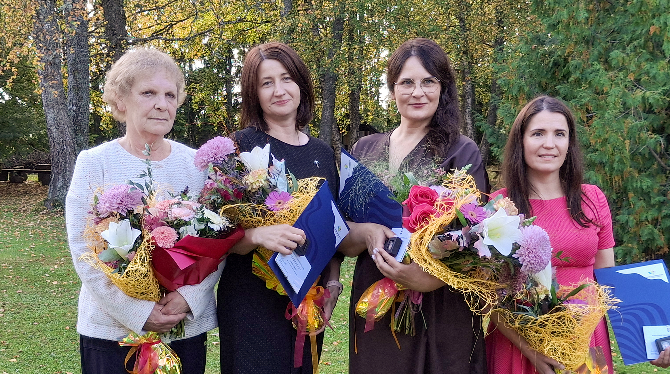 Neli naist seisavad sügiseste kollakate lehtedega puude taustal. Kõigil on käes lillekimbud. Vasakul on vanem halliseguste lühemate juustega naine heledas jakis, tema kõrval tumedate õlgadeni juustega tumesinises kleidis noorem naine, tema kõrval omakorda tumedate pikkade juustega naine tumepruunis kleidis ja kõige viimane tumedate pikkade juustega roosas kleidis lühem naine.
