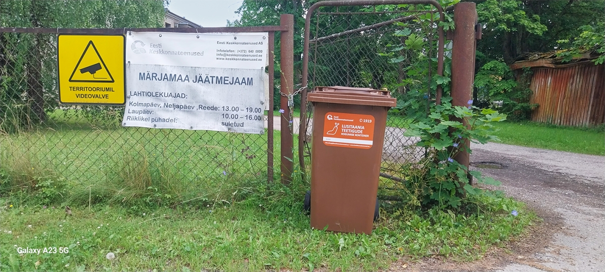 Roostes postidega metallaia ees seisab pruun konteiner, millel on oranž silt, kus vasakul pool valge teo pilt ja paremal valge tekst LUSITAANIA TEETIGUDE KOGUMISE KONTEINER. Konteinerist paremal on avatud värav, millest viib sisse asfalttee. Konteinerist vasakul on võrkaiale paigaldatud valge silt, millel mustas kirjas Märjamaa jäätmejaama lahtiolekuajad.