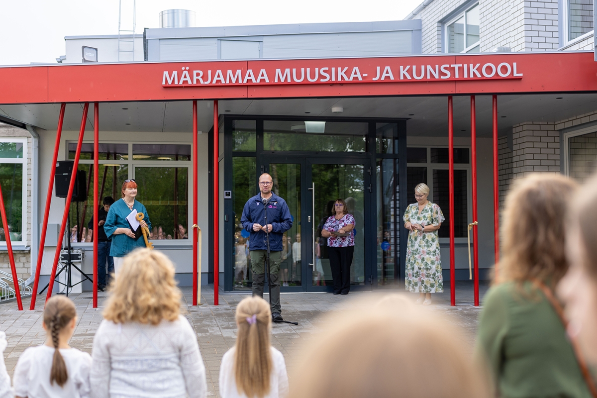 Esiplaanil seisavad lapsed seljaga vaataja poole. Tagapool on hoone sissepääs klaasidest ustega, mille ees keskel seisab ja peab kõnes sinises jopes ja rohekates pükstes mees. Temast paremal seisab blondide lühikeste juustega naine, kel on seljas rohekate, roosade ja kollaste roosidega kleit. Pildil vasakul seisab punaste tahapoole kinniseotud juustega naine, kel on seljas sinine pluus ja heledad püksid. Hoone katuse esiäär on punane ja sellel on valge kiri: MÄRJAMAA MUUSIKA- JA KUNSTIKOOL.