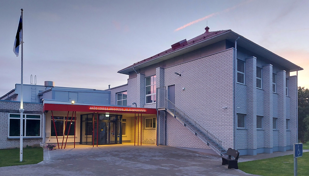 Paremal hallidest telliskibvides kahekordne hoone, mille esiküljel on metallist trepp. Paremal on madalam hoone osa, kus klaasist valgustatud sissepääsu kohal on katus ja selle alumisel punasel äärel on valge valgustatud silt MÄRJAMAA MUUSIKA- JA KUNSTIKOOL. Pilt on tehtud õhtul, taevas paremal on näha roosakat pilvekest.