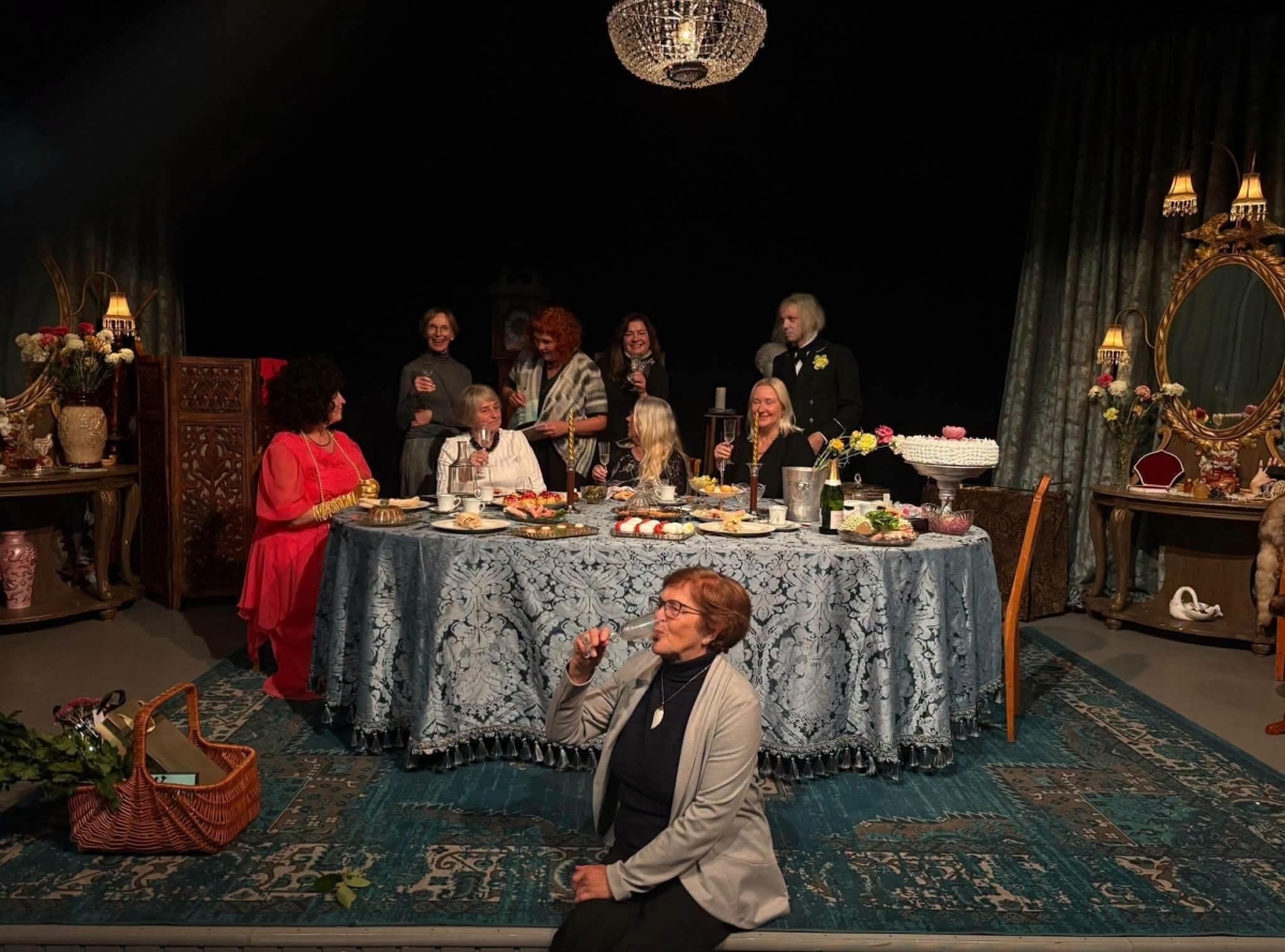 Teatrilaval on kaetud peolaua ümber grupp inimesi. Esiplaanil istub naine, pokaal käes.